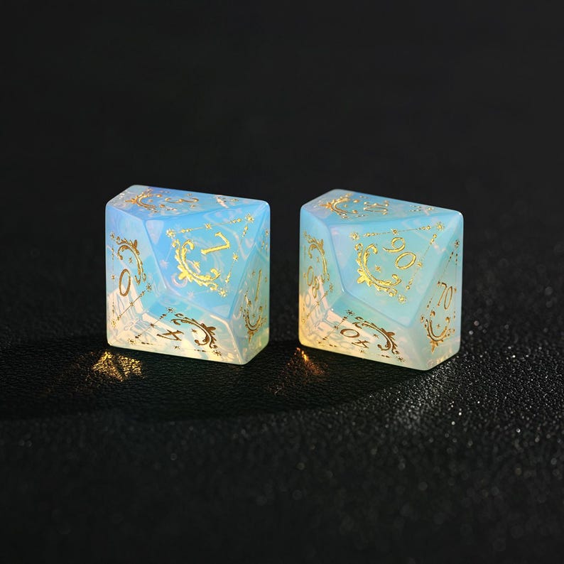 May include: Two opalescent dice with gold-colored filigree and numbers. The dice are a light blue color with a pearlescent sheen. The numbers are in a gold color. The dice are for a tabletop game.