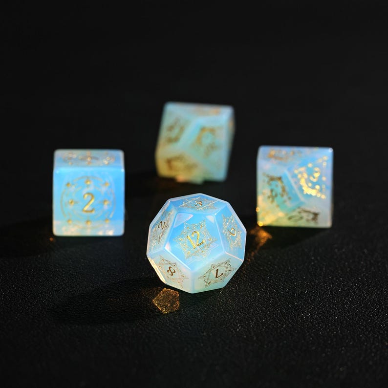 May include: Four dice, including a 20-sided die and three six-sided dice, are displayed. The dice are a translucent blue with gold-colored numbers and decorative accents. The dice are arranged on a dark surface, with the 20-sided die in the foreground.