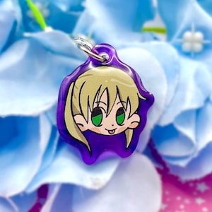 Op de afbeelding: Een paarse anime-stijl bedel met een zilveren sluiting. De bedel heeft een stripfiguur met blond haar, groene ogen en een glimlachende uitdrukking. De achtergrond is een cluster van blauwe bloemen.