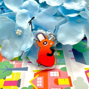 May include: A clear acrylic keychain featuring a cartoon character with an orange body, black arms, and a chainsaw head. The keychain is attached to a silver metal ring and black string. The background includes blue flower petals and a colorful house pattern.