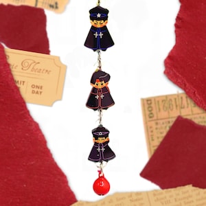 May include: A vertical charm featuring three small, stylized figures wearing dark purple capes and hats, connected by silver-toned metal jump rings. The top and bottom figures wear peaked caps with a white star, while a small, bright red bell hangs at the base.