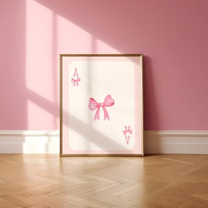Puede incluir: Impresión enmarcada de la carta de juego As de Lazos. La tarjeta presenta un fondo rosa claro y un borde rosa. Una cinta rosa está en el centro, con lazos más pequeños en las esquinas superior izquierda e inferior derecha. El marco es dorado.
