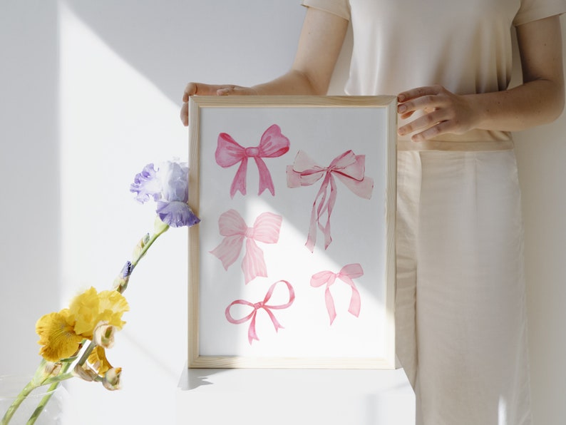 May include: A framed print with a white background featuring six pink watercolor bows of varying sizes and shapes.