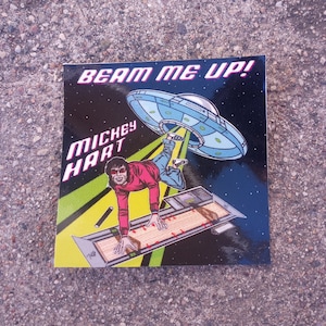 May include: A sticker with a cartoon image of a man in a red shirt and blue jeans being beamed up by a flying saucer. The text "Beam me up!" is at the top of the sticker and "Mickey Hart" is at the bottom.