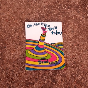 May include: A colorful sticker with the text "Oh, the trips you'll take!" and "with Dead and Co." A pink bear-like figure is at the top of a striped, cone-shaped structure. The background features a rainbow swirl design.