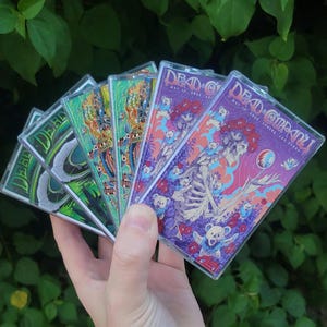 May include: A hand holds a fan of cassette tapes with colorful artwork. The tapes feature the words "Dead & Company" and various designs, including skeletons, bears, and floral patterns. The background is a lush green foliage.