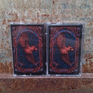 May include: Two identical cassette tapes in clear plastic cases featuring a red cardinal bird illustration centered within an ornate red and blue frame. The artwork includes intricate swirling patterns and the text July 14 2025 at the bottom.