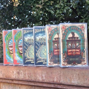 May include: A collection of six cassette tapes featuring colorful artwork. The tapes display the text "DEAD & COMPANY" and "CELEBRATING 60 YEARS". The artwork includes images of a cable car, cityscapes, and floral designs. The tapes are arranged in a row.