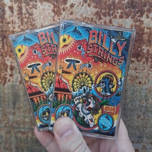 May include: Two clear plastic cassette cases featuring colorful artwork with the text Billy Strings. The design includes a sun, a ferris wheel, a circus tent, a creature, and a person with a guitar, all set against a vibrant orange and blue sky.