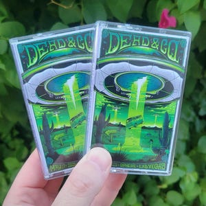 May include: Two cassette tapes with a vibrant green and black design. The artwork features a UFO, a desert landscape with cacti, and the text "DEAD & CO". Additional text includes "MAY 17 2025" and "025-SPHERE-LAS VEGAS".