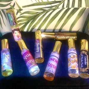 May include: A collection of eight small glass bottles with gold caps and labels. The labels have different designs and text, including "Kiss", "Sultry", "In the Rose", "Oh, hee-ha", "Lourdes of Hyde Park", "Australiana", and "Boque Ions".