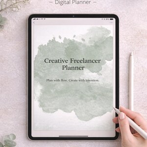 以下が含まれることがあります： 「Creative Freelancer Planner」のテキストが表示されたタブレットのデジタルプランナー。画面には、緑色の水彩画風のデザインが表示されています。スタイラスが画面の近くに保持されています。背景は明るくニュートラルな色です。