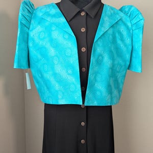 May include: A teal bolero jacket with a swirling pattern over a black collared shirt and button-down dress. The jacket has short sleeves and puffed shoulders. The dress has a row of buttons down the front.
