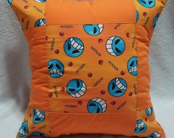 May include: Orange pillow with a blue and white pattern of smiling faces. The pillow is made of fabric and has a square shape.