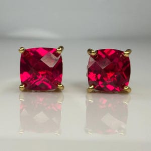 May include: A pair of stud earrings featuring square-cut, vibrant red gemstones. Each stone is faceted to catch the light and is securely held in place by four gold-toned metal prongs at the corners, creating a classic and symmetrical appearance.