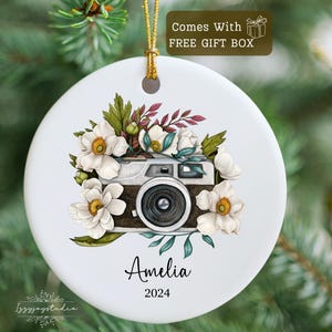 May include: White ceramic Christmas ornament with a vintage camera and white flowers. The ornament is personalized with the name "Amelia" and the year "2024".