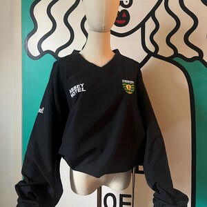 May include: Black long-sleeved pullover with a V-neck. The shirt features the text "ABBEY HOTEL" in white on the left chest and a crest on the right chest. The sleeves are gathered at the wrists. The shirt is displayed on a mannequin.