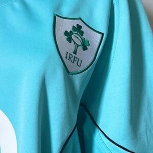 May include: A light blue rugby jersey with a black collar and trim. The jersey features a shield-shaped emblem with a green shamrock and the letters "IRFU". The number "9" is visible on the jersey.