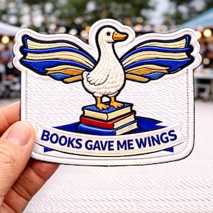 May include: Embroidered patch featuring a white goose with blue and gold wings, standing on a stack of books. The patch has the text "BOOKS GAVE ME WINGS" on a blue banner. The patch is rectangular with a white background.
