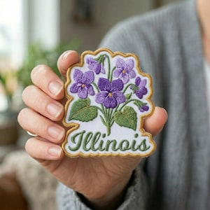 May include: Embroidered patch featuring a cluster of purple violets with green leaves and stems. The patch has a gold border and the word "Illinois" in green script.