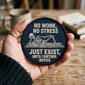 May include: A navy blue round patch with white embroidered text and a skeleton illustration. The text reads "NO WORK, NO STRESS" and "JUST EXIST, UNTIL FURTHER NOTICE". The skeleton is lounging on a rug.