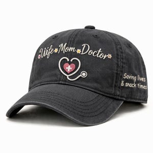May include: A dark gray baseball cap with embroidered text and graphics. The text reads "Wife Mom Doctor" with a heart and stethoscope graphic. Additional text on the side says "Saving lives & snack times".