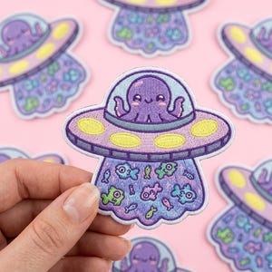 May include: Embroidered patch featuring a purple octopus piloting a UFO. The UFO is lavender with yellow accents and a light blue bubble. The bottom of the UFO has a design of colorful fish. The patch is held by a hand.