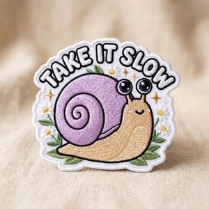 May include: Embroidered patch featuring a cartoon snail with a purple shell and a tan body, surrounded by daisies and green leaves. The patch has the text "TAKE IT SLOW" in white letters on a black border. A cute and whimsical design.