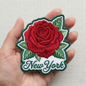 May include: Embroidered patch featuring a vibrant red rose with green leaves and a white background. The patch is outlined in green and has the words "New York" in a cursive font.