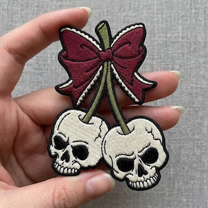 May include: Embroidered patch featuring two skulls as cherries, connected by green stems and topped with a burgundy bow. The skulls are off-white with black eye sockets and noses, outlined in black. The bow has a scalloped edge.