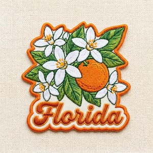 May include: Embroidered patch with white orange blossoms, green leaves, and an orange fruit. The patch has an orange border and the word "Florida" in an orange script font.