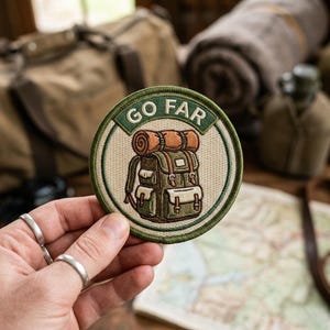 May include: Embroidered patch with a circular design featuring a green and brown backpack with a rolled-up sleeping bag on top. The patch has the words "GO FAR" in a green border. The patch is held by a hand with silver rings.