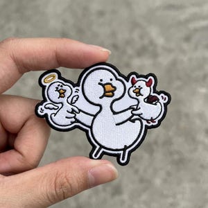 May include: Embroidered patch featuring a large white duck holding two smaller duck figures, one with a halo and the other with devil horns. The patch has a black border and is held up by a hand.