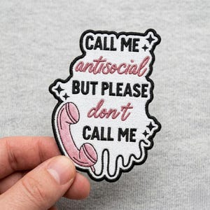 May include: Embroidered patch with the text "CALL ME antisocial BUT PLEASE don't CALL ME" in black and pink lettering. The patch features a pink telephone receiver graphic and a black border. The patch is held by a hand.