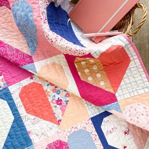 May include: A pink, blue, and red patchwork quilt with a heart pattern. The quilt is folded over a pink notebook with the word "NOTES" on the cover.