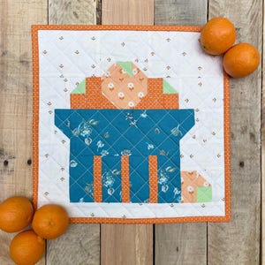 May include: A quilted wall hanging with a gift box design in teal, orange, and white. The box has vertical orange stripes and a floral pattern. Oranges are scattered around the wall hanging on a wooden surface.