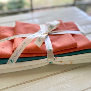 May include: A stack of fabric bundles in coral, teal, and white, tied with a white ribbon printed with "Heart Lake Quilts". The coral fabric has a stitched pattern. The white fabric has a floral design.