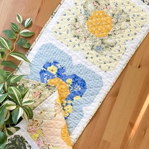 May include: A table runner with a floral pattern in shades of yellow, blue, green, and white. The runner is made of fabric and has a quilted design.