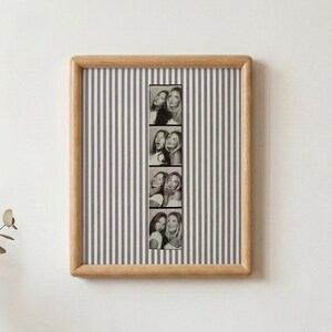 May include: A square wooden picture frame containing a vertical strip of four black and white photo booth portraits of two individuals. The background inside the frame features a pattern of thin, vertical grey and white stripes.