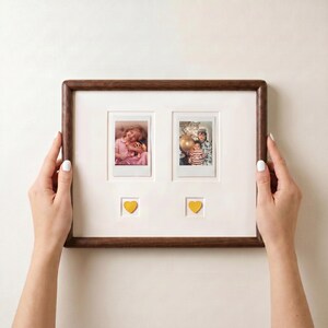 Op de afbeelding: Een houten fotolijst met twee kleine foto's en twee gele hartaccenten. De lijst heeft een donkerbruine afwerking en een witte passe-partout. De foto's tonen mensen. De lijst wordt door twee handen vastgehouden.