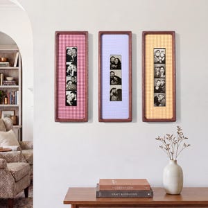 May include: Three vertical rectangular wall frames with dark wood borders, each containing a gingham fabric mat in red, light purple, or orange. Each frame displays a vertical strip of four black and white photo booth portraits of people.
