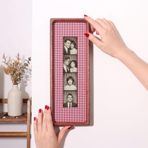 May include: A rectangular wooden frame containing a vertical strip of four black and white vintage photo booth portraits. The background inside the frame features a red and white gingham pattern. The frame is held against a plain white wall.
