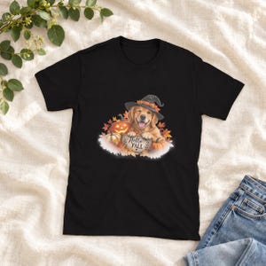 Può includere: Una t-shirt nera a maniche corte con la grafica di un golden retriever che indossa un cappello da strega, circondato da zucche e foglie autunnali. Il cane poggia le zampe su un cartello con la scritta Hello FALL e un piccolo cuore.