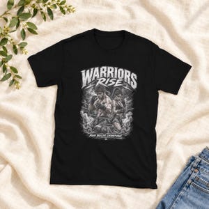 Può includere: Una t-shirt nera a maniche corte con una stampa grafica di due combattenti in un ring. Il testo recita WARRIORS RISE in alto e PAIN BUILDS CHAMPIONS in basso. Il design è realizzato in tonalità di grigio e bianco con effetti di fumo.