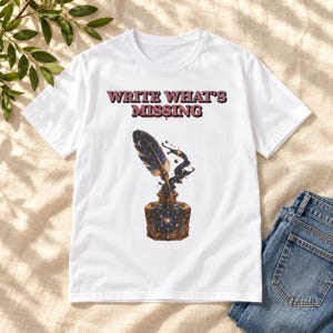 Può includere: Una t-shirt bianca con il testo "WRITE WHAT'S MISSING" in lettere rosso scuro sopra la grafica di una penna d'oca che emerge da un calamaio. Il calamaio e la penna sono decorati con motivi celesti, tra cui stelle, lune e una galassia.