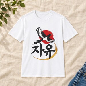 Puede incluir: Una camiseta blanca de manga corta con un estampado gráfico de un pájaro blanco y negro en vuelo sobre un fondo circular rojo. Debajo del pájaro, el texto coreano 자유 está impreso en negro, acentuado por un diseño de pincelada dorada curva.