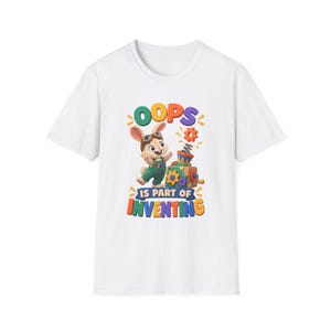 Könnte beinhalten: Ein weißes T-Shirt mit kurzen Ärmeln und einer Grafik eines Cartoon-Hasen, der eine Schutzbrille und eine grüne Latzhose trägt, neben einer mechanischen Spieluhr. Der Text lautet OOPS IS PART OF INVENTING in bunten, fetten Buchstaben.