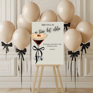 May include: A sign on an easel with the text "Brenda is a tini bit older" and a cocktail illustration. Beige balloons with black bows surround the sign. The sign welcomes guests to Brenda's 24th birthday party on 2.28.2025.