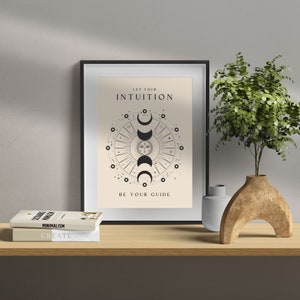 May include: A framed print with a beige background and black text that reads "Let your intuition be your guide". The print features a black and white illustration of a sun and moon with a circle of stars around it.