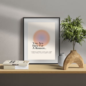 Op de afbeelding: Een ingelijst afdruk met een wazige oranje en blauwe cirkel in het midden. De tekst "You Are Here For A Reason..." staat onder de cirkel. De tekst gaat verder met "TO UNDERSTAND / TO EXPERIENCE / TO LEARN / TO FEEL / TO INSPIRE / TO LOVE / TO TEACH / TO TRY / TO CHANGE / TO REPLACT / TO HELP / TO CONNECT / TO CREATE"